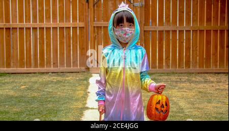 A kindergartener wearing a facemask holding a pumpkin jack-o-lantern during halloween during a pandemic. Concept covid 19 during halloween. Stock Photo