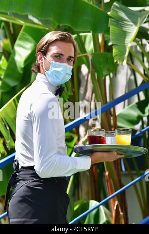 Close up of a young male waiter using a surgical mask holding a bar tray with assorted drinks while looking at the camera. Serving and safety concept. Stock Photo