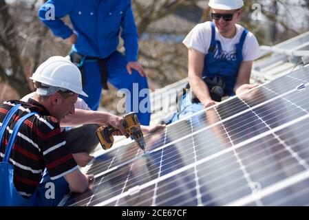 Male engineers installing solar photovoltaic panel system using screwdriver. Electricians mounting blue solar module on roof of modern house. Alternative renewable energy concept. Stock Photo