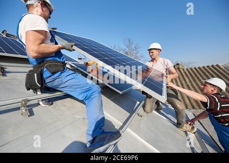 Male team engineers installing stand-alone solar photovoltaic panel system. Electricians lifting blue solar module on roof of modern house. Alternative energy ecological concept. Stock Photo