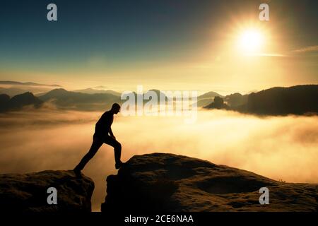 Crazy hiker in black is jumping between the rocky peaks. Wonderful daybreak in rocky mountains, heavy mist in deep valley. Mirac Stock Photo