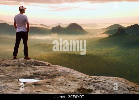 Tall man in white shirt and  black trousers with red baseball cap  stay on sharp cliff and watch to valley. Colorful  misty morn Stock Photo