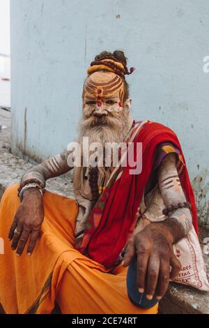 India - March 9, 2020: Aged Indian male with painted face and body and in traditional clothes relaxing on stone ground and looking at camera Stock Photo