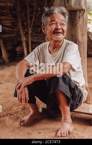Thailand - August 16, 2010: Cheerful senior Thai male in dirty clothes relaxing on sandy ground in countryside and looking away Stock Photo
