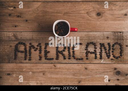 Top view composition of red mug with roasted aromatic coffee placed on wooden table above inscription americano made with coffee beans Stock Photo