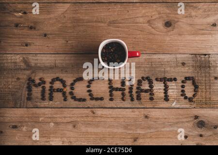 Top view composition of red mug with roasted aromatic coffee placed on wooden table above inscription macchiato made with coffee beans Stock Photo