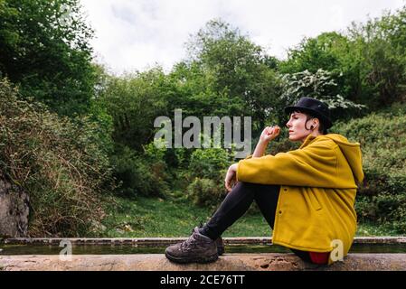 Side view of androgynous female tourist sitting on stone border of pond and enjoying vacation in Asturias Stock Photo