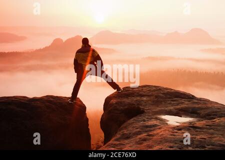 Hiker in black is jumping between the rocky peaks. Wonderful daybreak in rocky mountains, heavy orange mist in deep valley. Mira Stock Photo