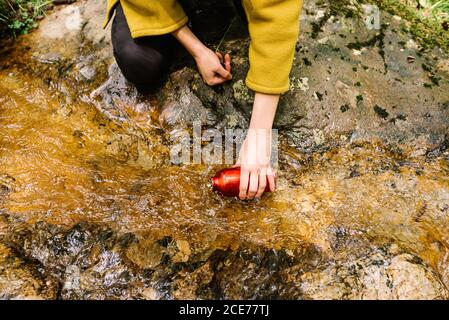 From above crop unrecognizable thirsty female in outerwear taking clean water from creek while resting in green forest Stock Photo