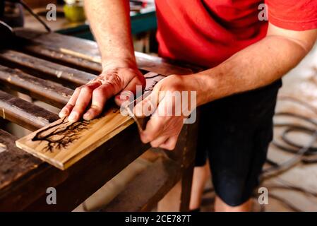 faceless male woodworker standing at workbench and polishing timber board with sandpaper while working in workshop Stock Photo
