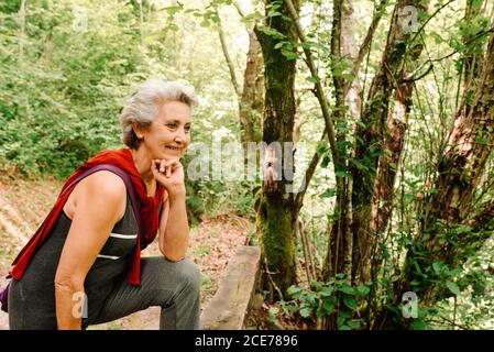 Side view of cheerful senior female traveler standing in woods and enjoying amazing landscape Stock Photo