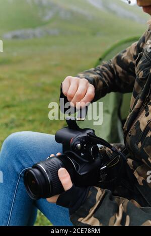 Crop anonymous male photographer in jeans and khaki jacket inserting memory card into photo camera while working in nature Stock Photo