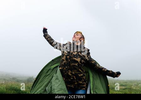 Positive female in warm clothes getting out of camping tent and stretching body during foggy morning in nature Stock Photo