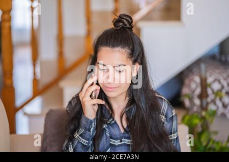 Young female freelancer in casual outfit sitting on sofa with laptop on knees and talking on mobile phone while working on remote project at home Stock Photo