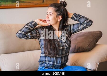 Smiling young female freelancer in casual outfit sitting on sofa in front of laptop and stretching while resting during work on project at home Stock Photo