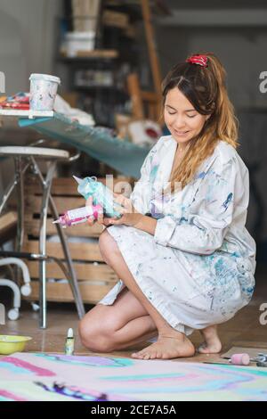 Smiling barefoot female in dirty robe squatting on wooden floor holding plastic bottle with paints while looking at abstract picture in art studio Stock Photo