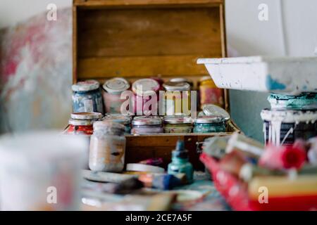 Open wooden box with collection of glass dirty jars with various paints surrounded by artist tools and paintbrushes in workshop Stock Photo