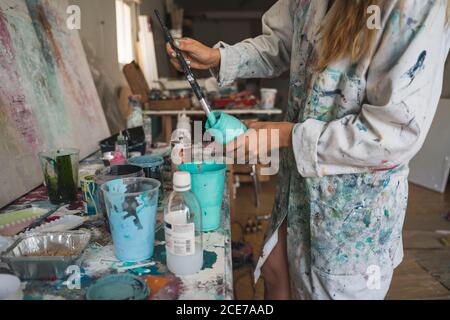 High angle side view of crop young female artist standing at counter with various tools and preparing colors for drawing while working in creative workshop Stock Photo