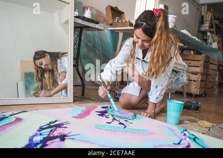 Cheerful barefoot female artist in dirty robe and gloves lying on wooden floor with closed eyes in front of abstract painting in art studio Stock Photo