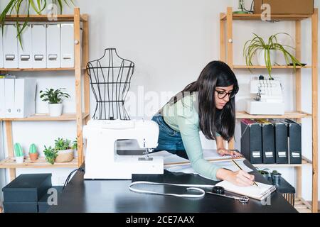 Young ethnic seamstress in eyeglasses taking notes standing leaned forward while using straightedge at table with papers and scissors in loft style studio Stock Photo