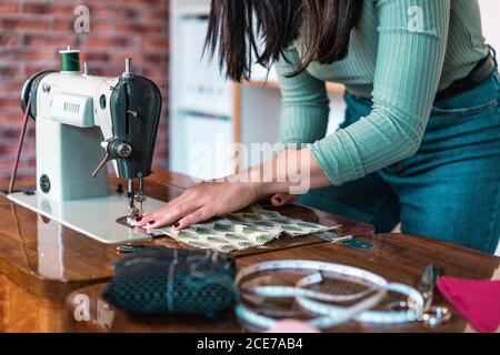 Crop unrecognizable slim female tailor in casual wear with manicure using sewing machine while standing leaned forward near table with soft fabric sample and measuring meter in loft style atelier Stock Photo