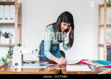 Ethnic female tailor in casual wear speaking on cellphone and taking note on sheet of paper while leaning forward near sewing machine and blank notepad with fabric samples in workshop Stock Photo