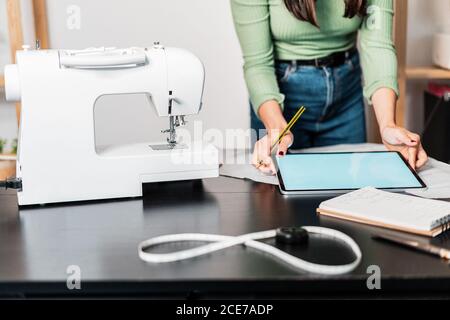 Crop unrecognizable seamstress standing leaned forward while using tablet and straightedge at table with papers in studio Stock Photo