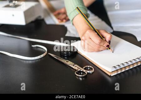 Young ethnic seamstress in eyeglasses taking notes standing leaned forward while using straightedge at table with papers and scissors in loft style studio Stock Photo