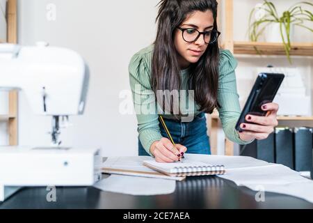 Young ethnic seamstress in eyeglasses taking notes standing leaned forward while using straightedge at table with papers and scissors in loft style studio Stock Photo