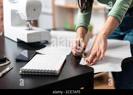 unrecognizable seamstress standing leaned forward while using straightedge at table with papers and scissors in loft style studio Stock Photo