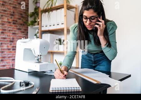 Young ethnic seamstress in eyeglasses taking notes standing leaned forward while using straightedge at table with papers and scissors in loft style studio Stock Photo