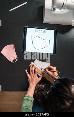 From above crop seamstress standing leaned forward while using tablet and straightedge at table with a mask draw with papers in studio Stock Photo