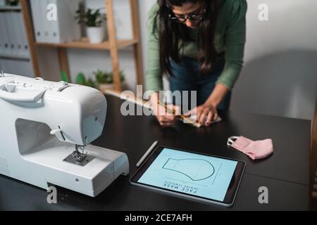 From above crop seamstress standing leaned forward while using tablet and straightedge at table with a mask draw with papers in studio Stock Photo