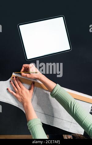 from above crop seamstress standing leaned forward while using straightedge at table with papers and tablet in studio Stock Photo