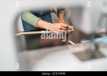 From above crop seamstress standing leaned forward while using tablet and straightedge at table with a mask draw with papers in studio Stock Photo