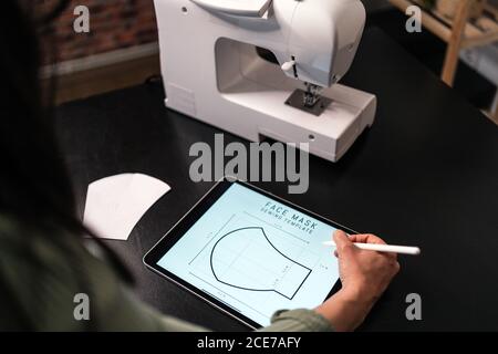 from above crop seamstress standing leaned forward while using tablet and straightedge at table with a mask draw with papers in studio Stock Photo