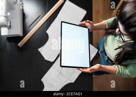from above crop seamstress standing leaned forward while using tablet and straightedge at table with papers in studio Stock Photo