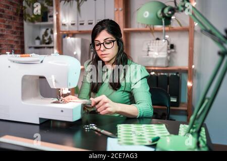 Slim female tailor in casual wear using sewing machine while sitting in table with sewing machine in loft style atelier Stock Photo