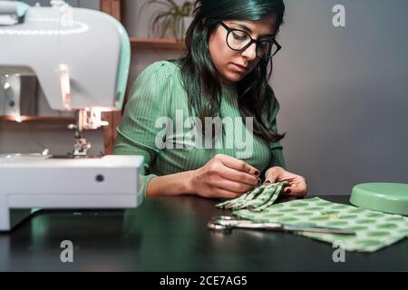 Young female tailor sitting in table using needles to stitch face masks near modern sewing machine in loft style atelier Stock Photo