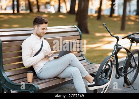 Work during break outdoors. Attractive guy sits on bench, types at laptop with coffee and bike Stock Photo