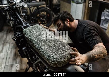 Tattooed male biker standing in shabby garage and working on seat while fixing motorbike Stock Photo