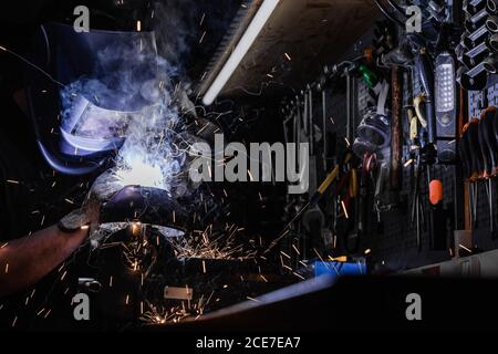 Side view of anonymous male worker in protective helmet standing at workbench and fixing metal details with welding tool Stock Photo