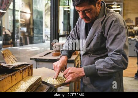 Concentrated middle aged ethnic goldsmith in uniform working with gold on wooden table using a pencil in studio Stock Photo