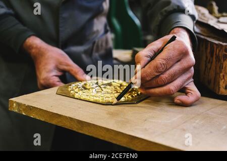 Crop of concentrated middle aged ethnic goldsmith in uniform working with gold on wooden table using a pencil in studio Stock Photo