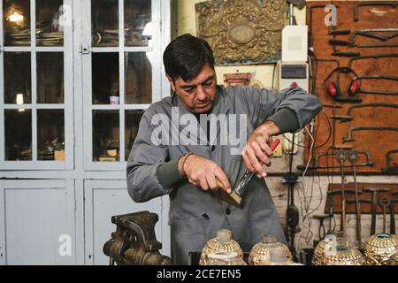 Focused middle aged ethnic craftsman in uniform using pincers while working with golden stick near various vases behind wall with tools in workshop Stock Photo