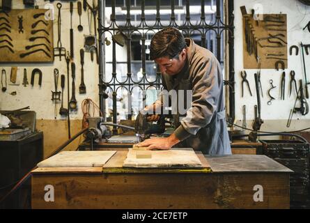Concentrated middle aged ethnic artisan in robe working with wooden blocks at table using drill while stranding near window with fence and wall with professional tools Stock Photo