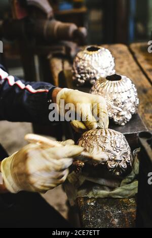 From above of crop anonymous craftsman in gloves sitting at old wooden table while cleaning gold vase with creative ornament using brush in studio Stock Photo