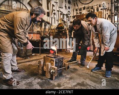 Group of professional male artisans working in weathered workshop and pouring molten metal into mold cavity during metal casting process Stock Photo