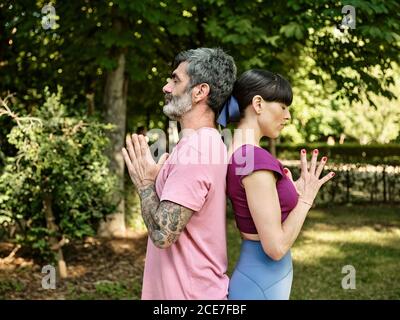 Side view of tranquil man and Woman practicing yoga in Tadasana while standing back to back with closed eyes and Namaste hands in park Stock Photo