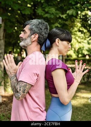 Side view of tranquil man and Woman practicing yoga in Tadasana while standing back to back with closed eyes and Namaste hands in park Stock Photo